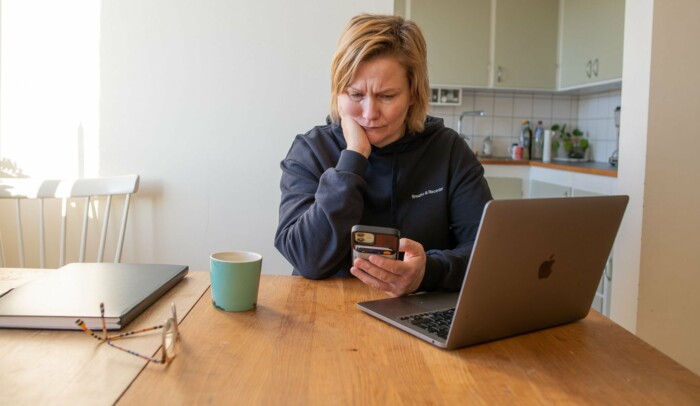 Kvinna sitter vid bord med sin mobil i handen och ser bekymrad ut